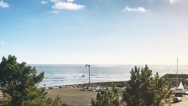 A coastal view with a clear sky, a rocky shoreline, and a parking area with a few parked cars. - Adorable T2 apartment (45m2) in a residence with panoramic sea view (Jard-sur-Mer)