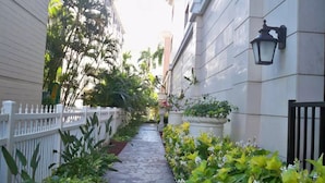 A narrow stone pathway flanked by lush greenery and white fences, leading to a building with a white facade and a black lantern-style light fixture. - Royal Gardens Waikiki - Sleeps 3 Full Kitchen  (Honolulu)
