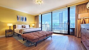 A hotel room with a large bed, a bench, a nightstand, a dresser, and a view of the city through large windows. - Royal Gardens Waikiki - Sleeps 3 Full Kitchen  (Honolulu)