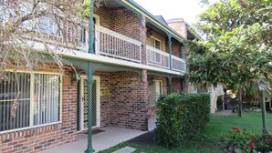 A two-story brick building with a balcony, a sign indicating holiday letting, and a well-maintained garden with red flowers.