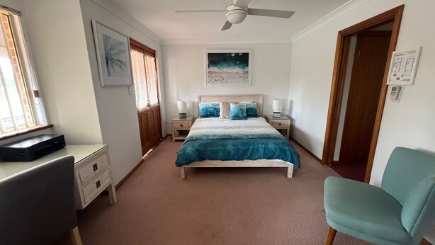 A bedroom with a bed, bedside tables, a desk, a chair, a ceiling fan, and a window with curtains. - Comfortable Townhouse with WIFI right in the heart of town. (South West Rocks)
