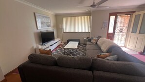 A living room with a sofa, a TV, a coffee table, and a window with blinds. - Comfortable Townhouse with WIFI right in the heart of town. (South West Rocks)