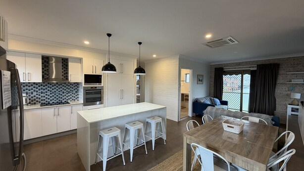 A modern kitchen with white cabinets, a central island with stools, a dining table, and a living area with a couch and large windows. - CAMEO COURT UNIT 9 - Beautiful Unit close to town centre. (South West Rocks)