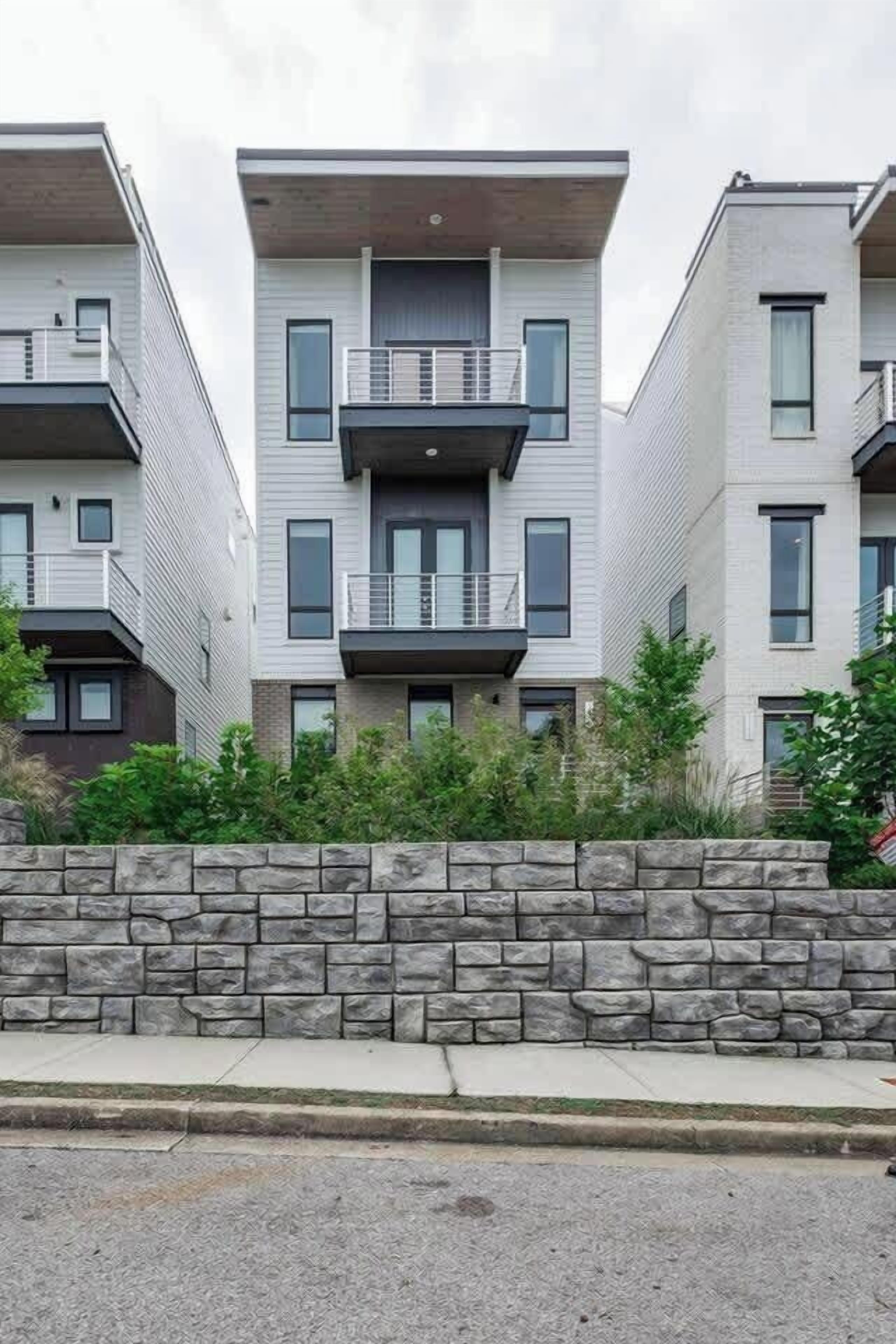 A modern multi-story residential building with balconies, a stone retaining wall, and a paved sidewalk.