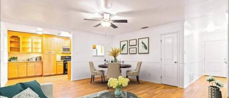 A spacious living area with a dining table, chairs, a ceiling fan, and a kitchen in the background.