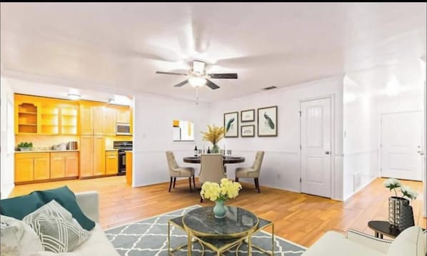 A spacious living area with a dining table, chairs, a ceiling fan, and a kitchen in the background.