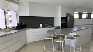 A modern kitchen with white cabinets, a granite countertop, and a built-in island with bar stools. - Sitka Penthouse Apartment (Maroochydore)
