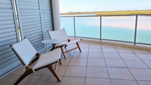 A balcony with two white wicker chairs and a small round table, overlooking a calm body of water and a distant shoreline. - Sitka Penthouse Apartment (Maroochydore)