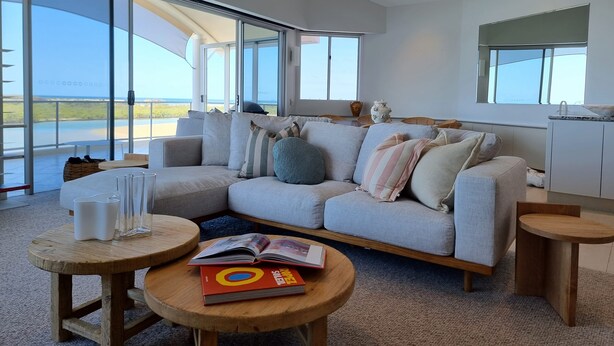 A modern living room with a large sectional sofa, a wooden coffee table, and a view of the ocean through large windows. - Sitka Penthouse Apartment (Maroochydore)