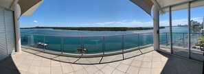 Een balkon met glazen hekwerk met uitzicht op een rustig wateroppervlak, een eiland in de verte en een stadsgezicht.
