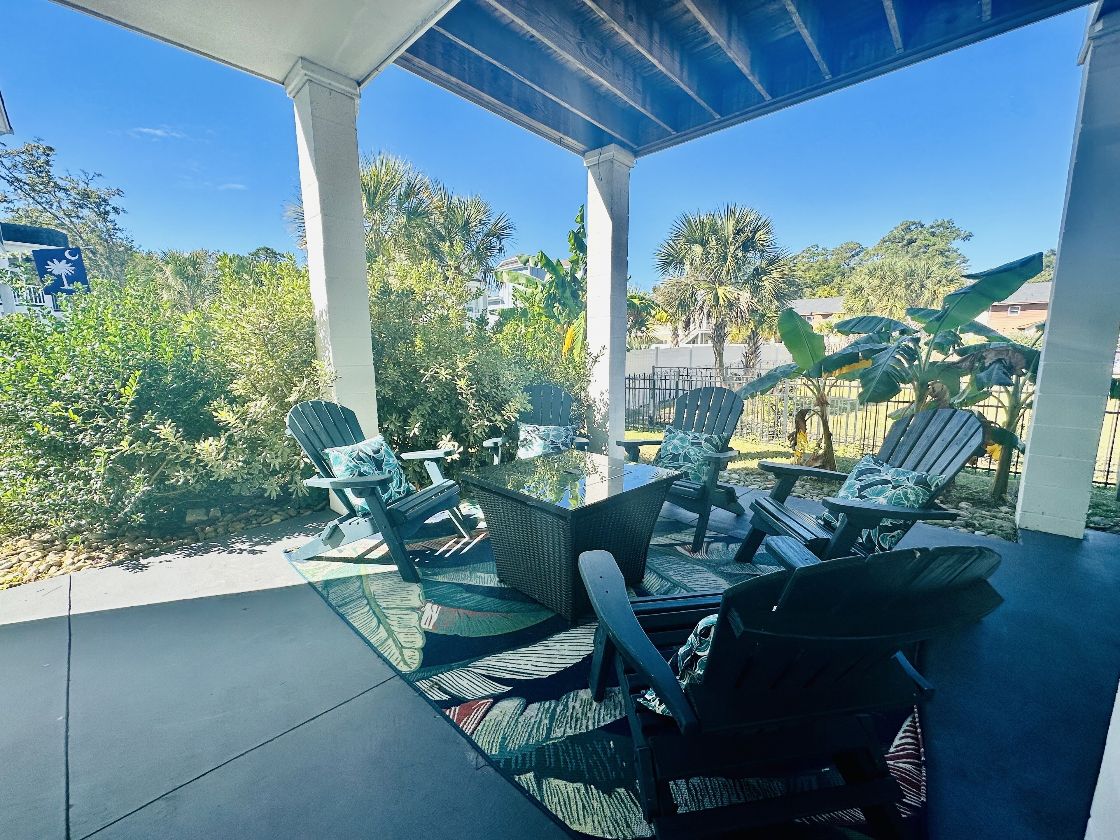 Un patio couvert aménagé avec du mobilier d’extérieur, notamment des chaises et une table, entouré d’une végétation luxuriante et de palmiers.
