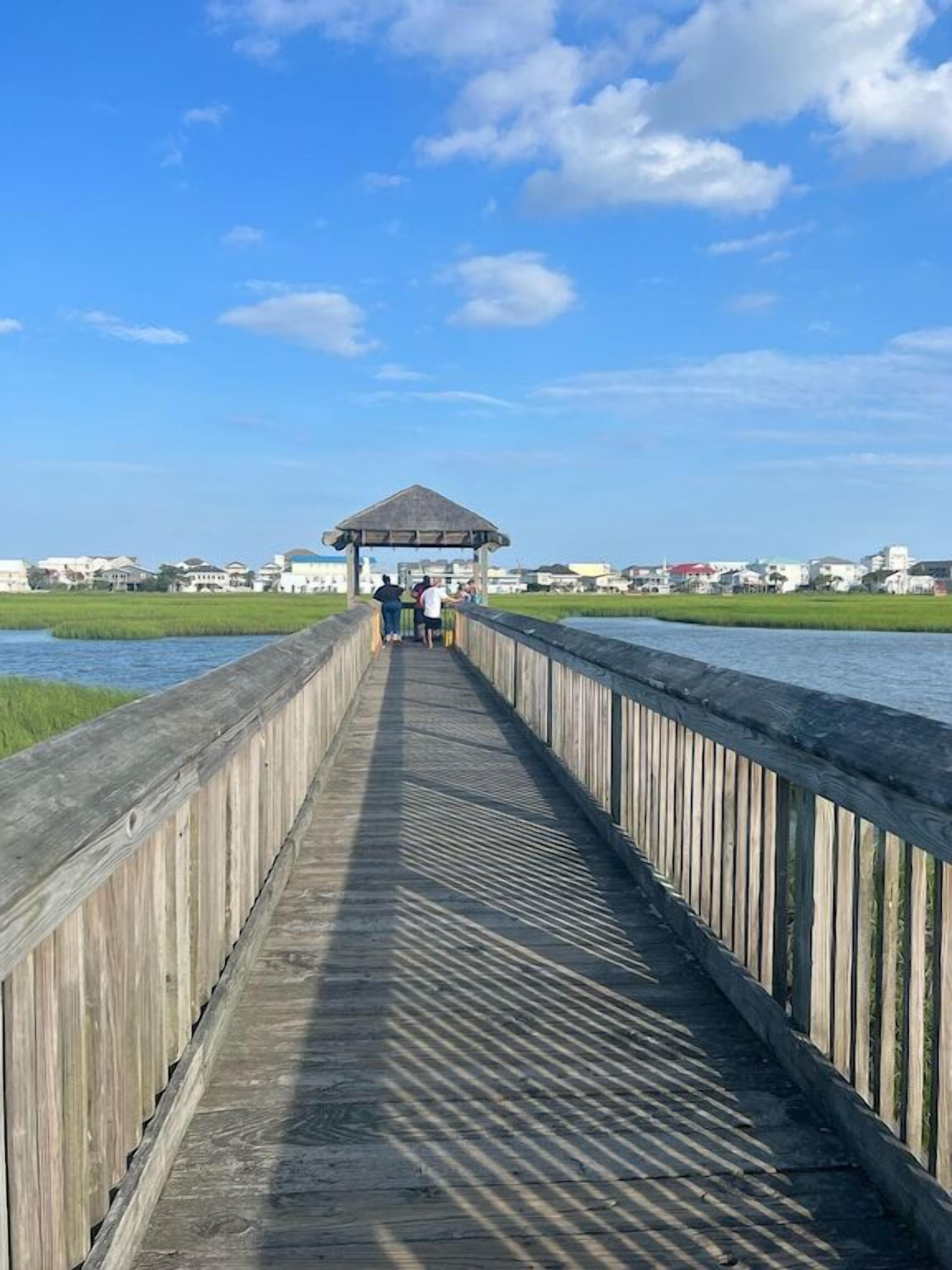 Une promenade en bois mène à un belvédère situé au-dessus de l’eau, avec des maisons en arrière-plan.