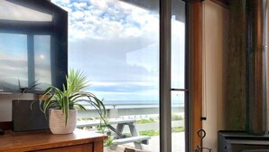A cozy living room with a large window overlooking a deck and the ocean, a fireplace, and a wooden table with a plant. - Surf Shack - Beach Front (Separation Creek)