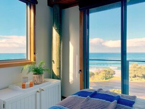 Een slaapkamer met een groot raam met uitzicht op de oceaan, een witte radiator en een nachtkastje met een plant en boeken.