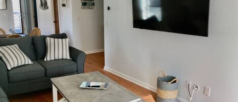 A living room with a grey sofa, striped pillows, a white coffee table, and a flat-screen TV mounted on the wall.