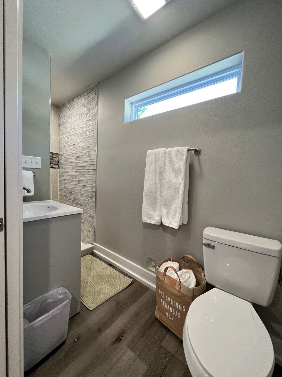 A bathroom with a white toilet, a sink, a towel rack, a small window, and a wooden floor.