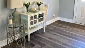 Een kamer met een houten vloer, een glazen tafel op metalen poten, een witte console met glazen deuren, een lamp, een schilderij en een deur met twee haakjes.