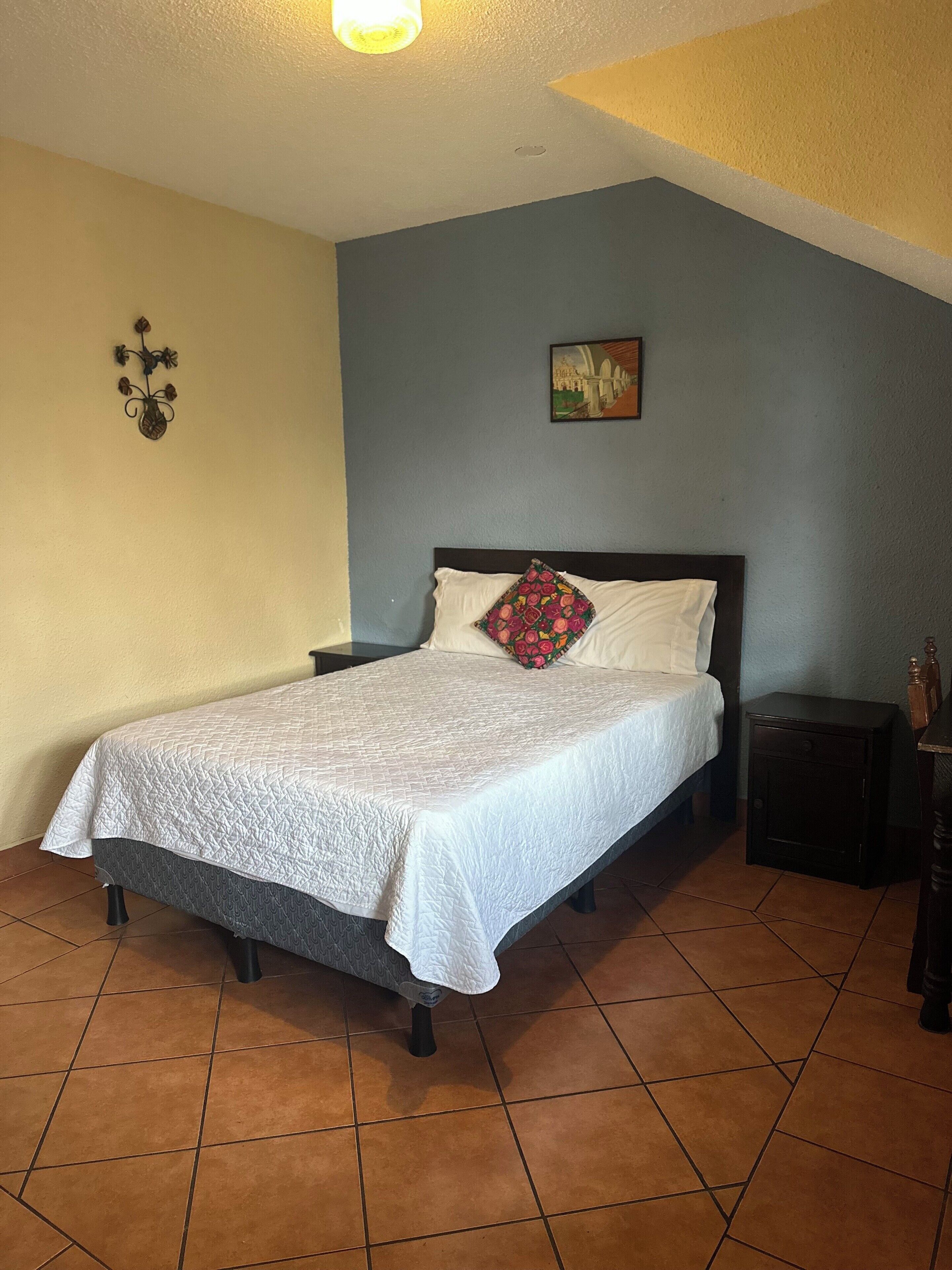 Una cama bien hecha con colcha blanca, un cojín decorativo y una mesita de noche con lámpara.