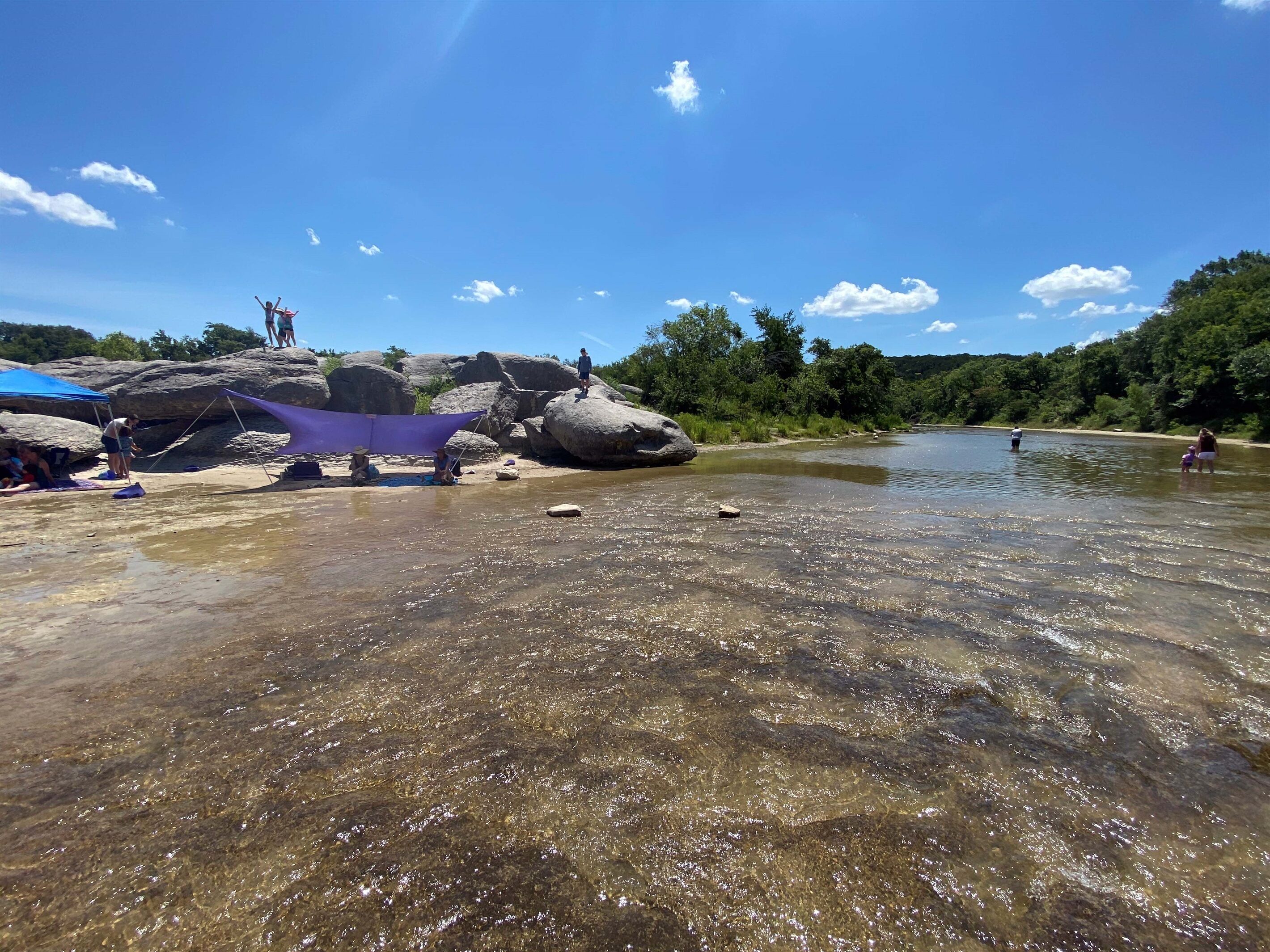 A riverbank with clear water, rocks, and people engaging in various activities.