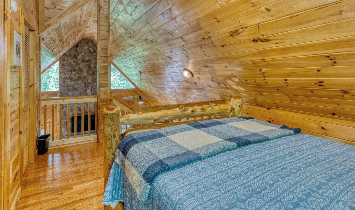 A wooden cabin bedroom with a checkered bedspread, a stone fireplace, and a balcony with a wooden railing.