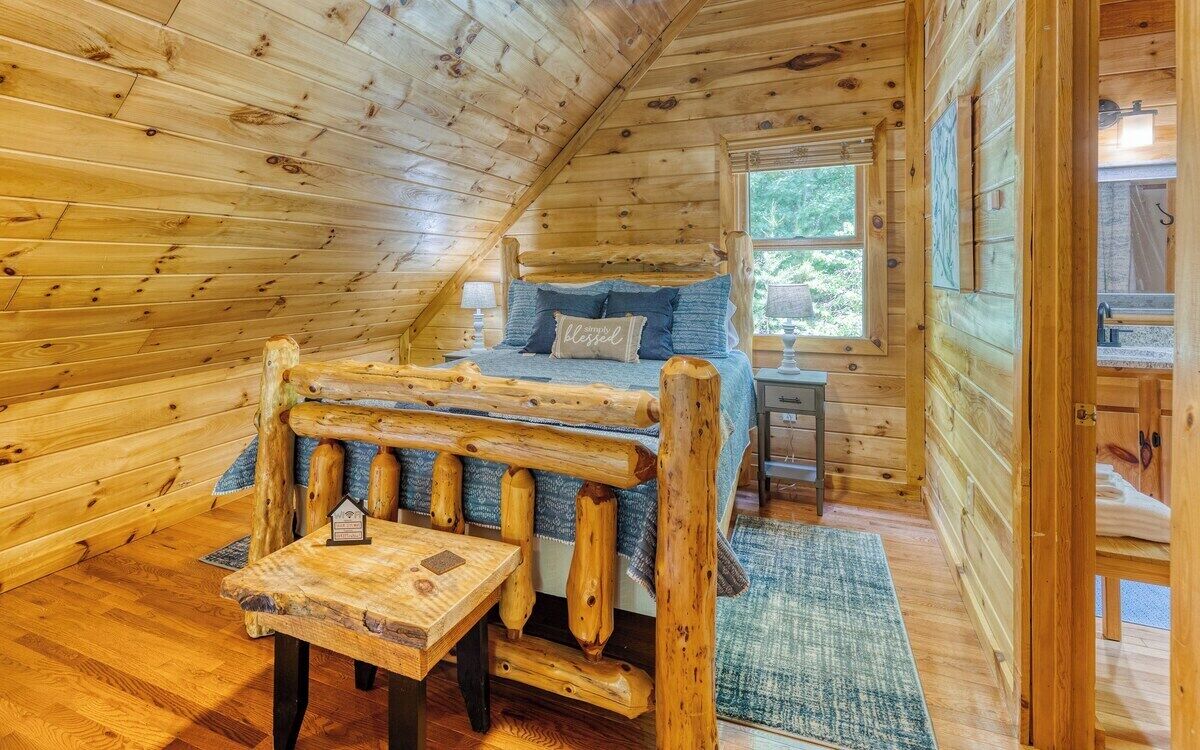 A cozy log cabin bedroom with a wooden bed, a small table, and a window providing a view of the outdoors.
