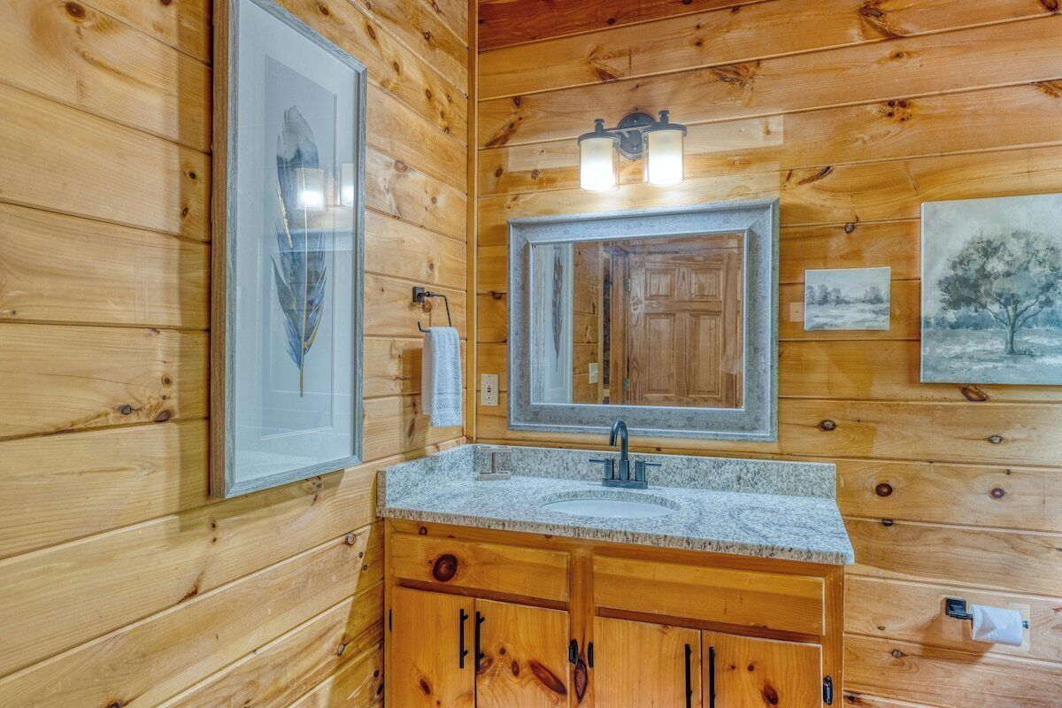 A wooden cabin bathroom with a granite countertop sink, wooden cabinets, a large mirror, and framed artwork on the walls.