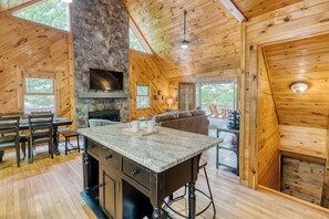 A cozy cabin interior with a stone fireplace, wooden walls, a dining area, and a large window overlooking a deck. - Mountain Cabin|Covered Deck|Hot Tub|Wifi|Gameroom (Morganton)