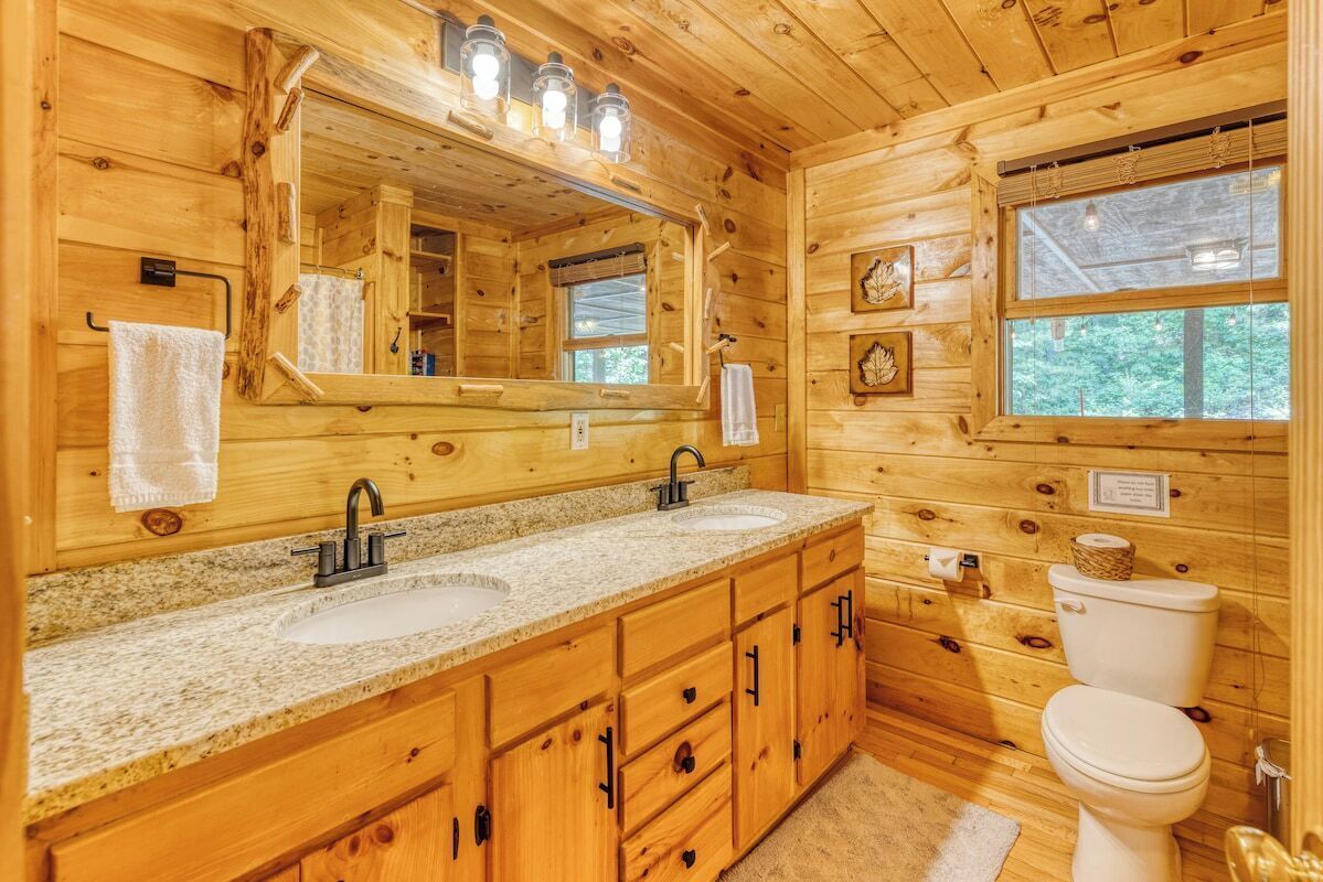 Une salle de bain rustique, avec des murs en bois, comprenant deux lavabos, un grand miroir et des toilettes.