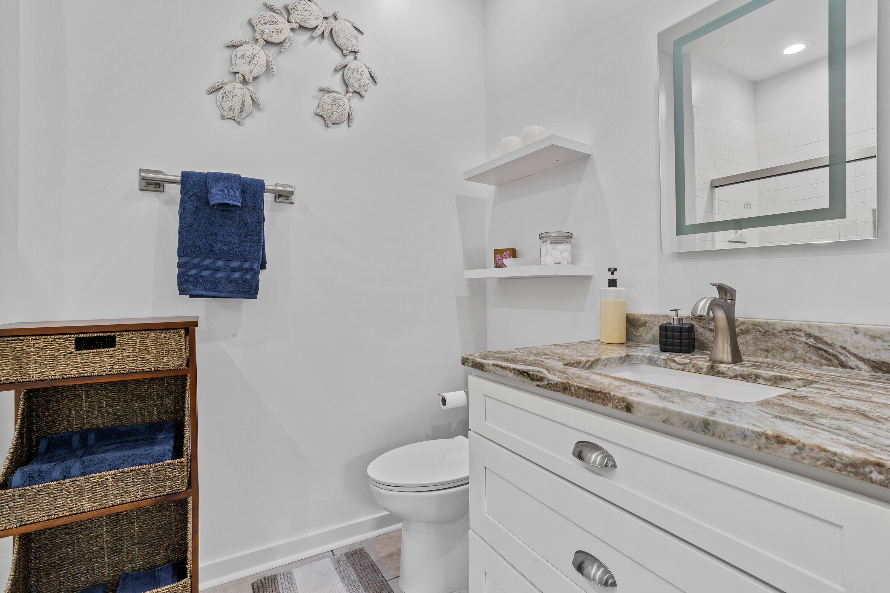 Un baño moderno con inodoro blanco, lavamanos de mármol y un espejo empotrado. Hay cestas de mimbre en un estante y una toalla azul colgada en un tendedero metálico.