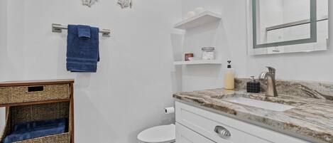 Un baño moderno con inodoro blanco, lavamanos de mármol y un espejo empotrado. Hay cestas de mimbre en un estante y una toalla azul colgada en un tendedero metálico.