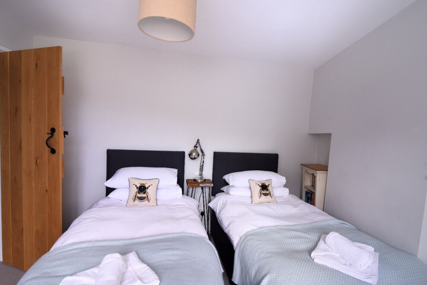 A bedroom with two beds, each with a pillow featuring a bee design, a bedside table with a lamp, and a wooden wardrobe.
