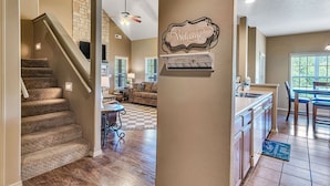 Un intérieur de maison avec un escalier menant à un espace de vie, une cuisine avec un bar et une pancarte « Bienvenue » accrochée au mur.