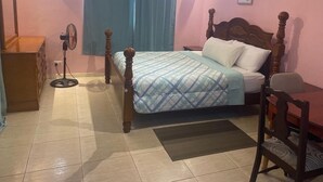Een slaapkamer met een bed, een bureau, een stoel, een dressoir, een ventilator en een raam met gordijnen.