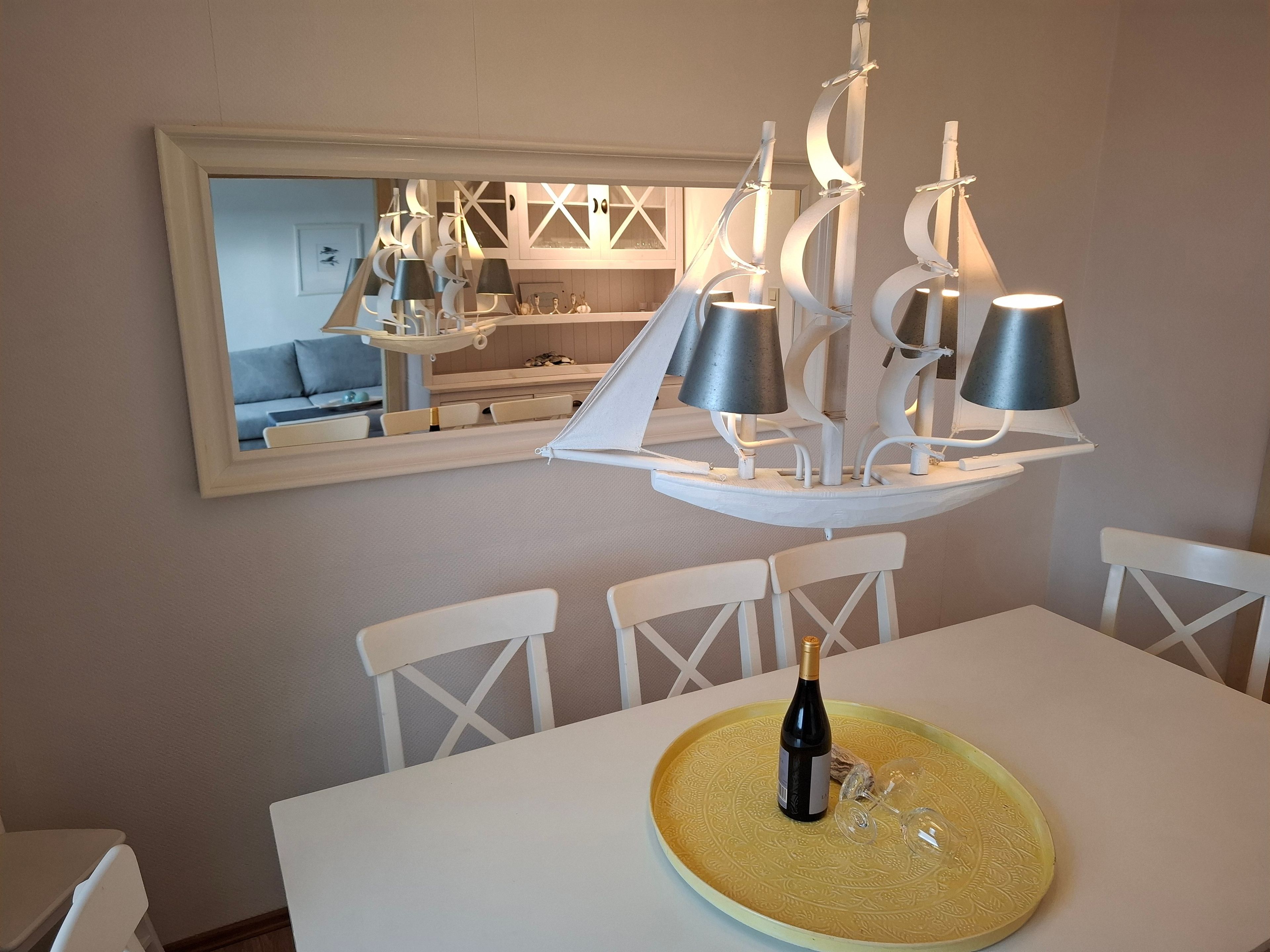 Un coin repas avec une table blanche, des chaises, un set de table jaune, une bouteille de vin et un lustre moderne.