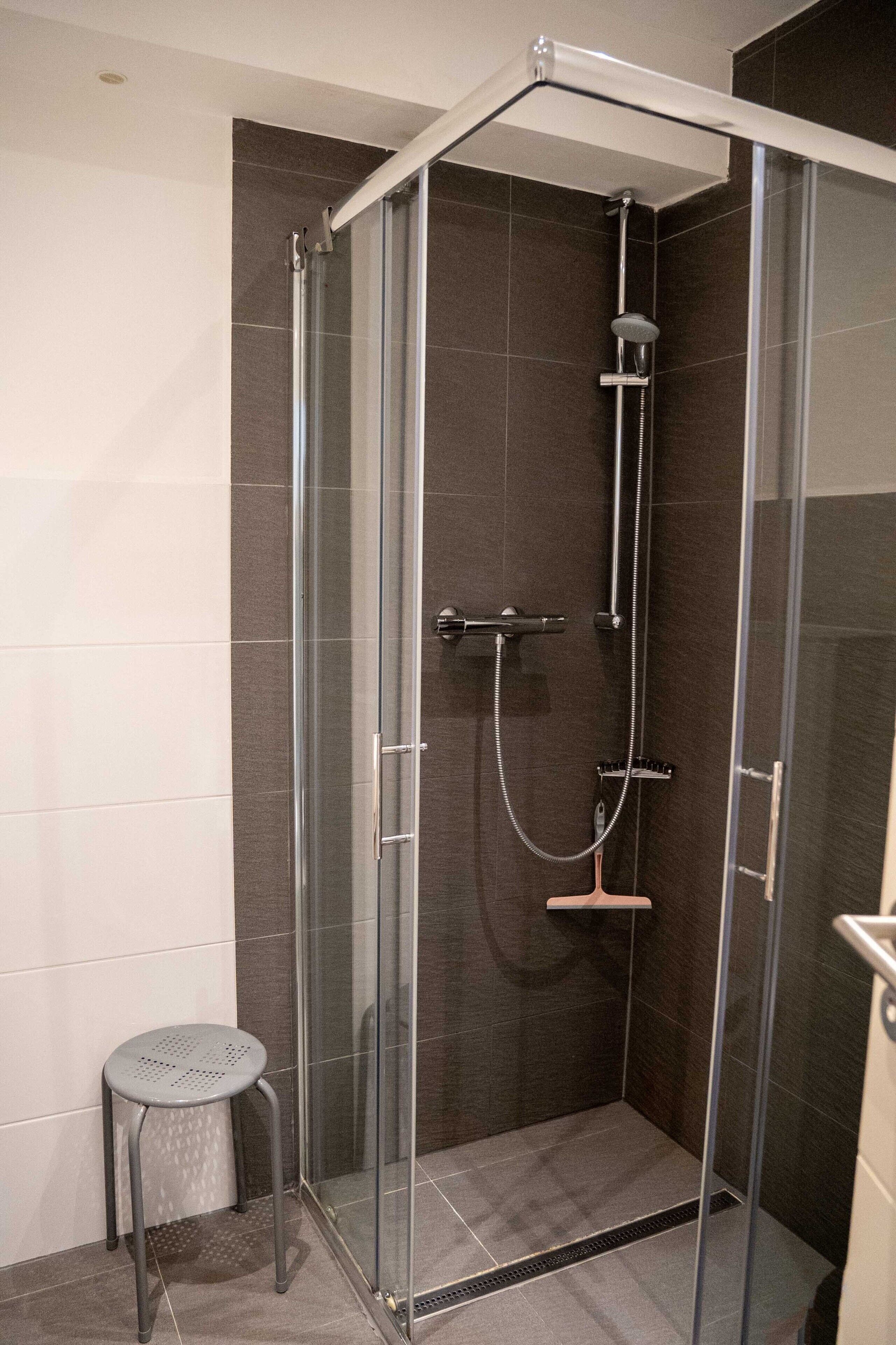 A glass-enclosed shower stall with a handheld showerhead, a handheld shower head, and a small stool.