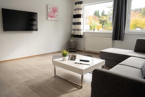 Ein modernes Wohnzimmer mit Flachbildfernseher, weißem Couchtisch, grauem Sofa, Topfpflanze und einem Fenster mit Vorhängen.