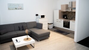 Una sala moderna con un sofá seccional gris, una mesa de centro blanca y una cocina con armarios blancos, un microondas y una estufa.
