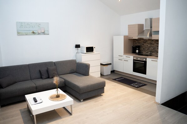 Un salon moderne avec un canapé d’angle gris, une table basse blanche et un coin cuisine doté de meubles blancs, d’un four à micro-ondes et d’une cuisinière.