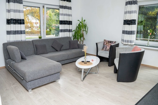 Ein modernes Wohnzimmer mit einem grauen Ecksofa, einem runden Couchtisch, einem schwarzen Sessel und einem kleinen Beistelltisch mit einer Vase.