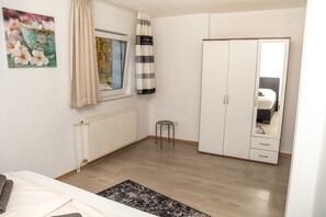 Ein Schlafzimmer mit einem Bett, einem Heizkörper, einem Fenster mit Vorhängen, einem kleinen Hocker und einem Kleiderschrank.