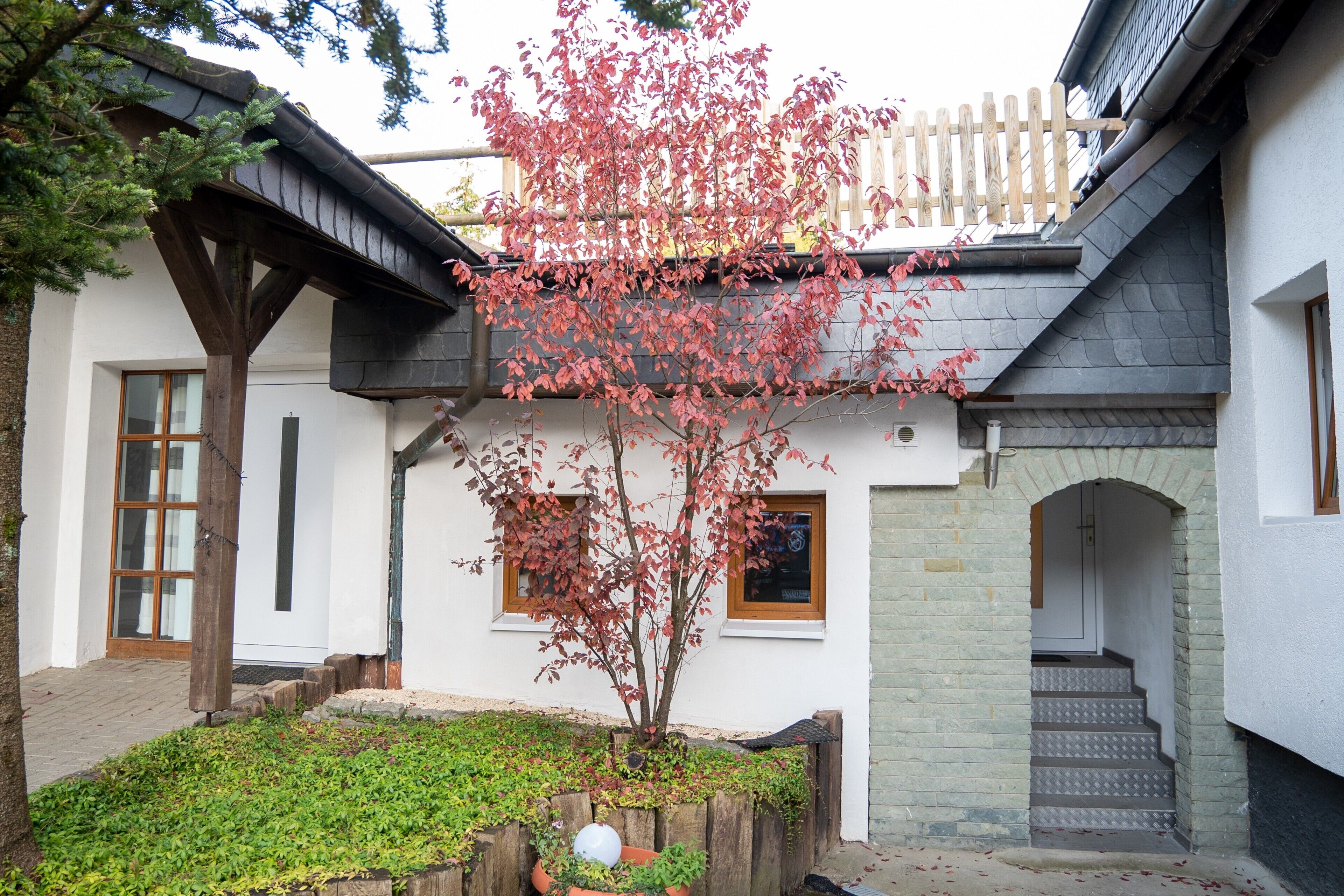 A building with a wooden staircase, a small garden with a tree, and a white exterior wall.