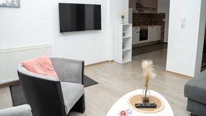A modern living room with a flat-screen TV, a grey sofa, a small table with a vase, and a kitchen in the background. - 'Villa Nordhang - Apartment 4' with Mountain View, Balcony & Wi-Fi (Winterberg)