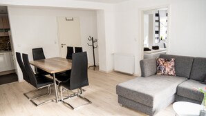A modern living room with a grey sofa, a dining table with chairs, and a coat rack.