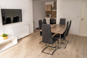 A modern dining area with a wooden table, black chairs, a TV mounted on the wall, and a kitchen in the background.