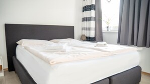 Un lit bien fait, avec une tête de lit grise, une table de chevet sur laquelle se trouve une lampe, et une fenêtre avec des rideaux.