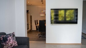 Un salón moderno con un sofá gris, un televisor de pantalla plana montado en la pared y un comedor al fondo.