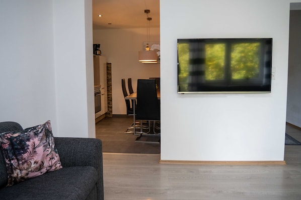 Un salon moderne avec un canapé gris, un téléviseur à écran plat fixé au mur et un coin repas à l’arrière-plan.