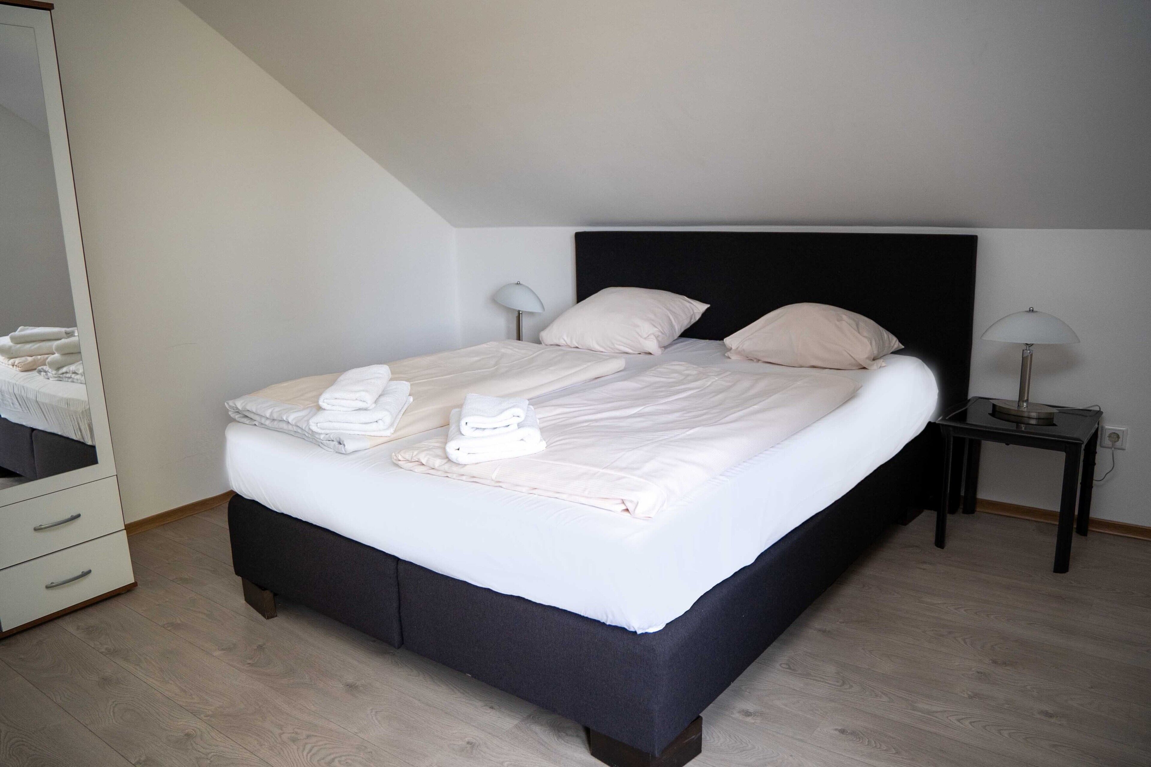 A neatly made bed with pillows and folded towels, a bedside table with a lamp, and a dresser with drawers.