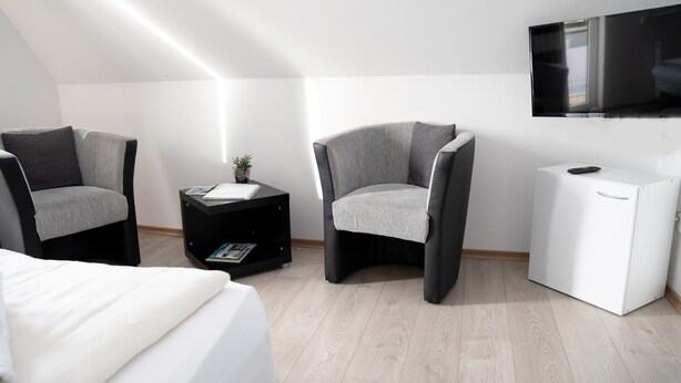 Ein modernes Zimmer mit einem Bett, zwei Sesseln, einem kleinen Tisch mit Pflanze, einem weißen Schrank und einem an der Wand befestigten Flachbildfernseher.