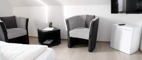 A modern room with a bed, two armchairs, a small table with a plant, a white cabinet, and a flat-screen TV mounted on the wall.